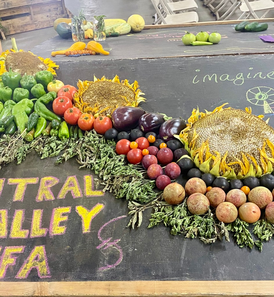 A colorful swoop of tomatoes, potatoes, peppers, greens, other vegetables, and sunflower heads, arranged on a chalkboard.